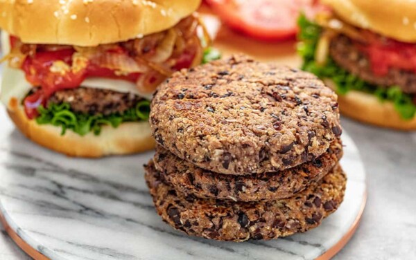 Three Black Bean Burger Patties stacked on a marble slab with a full burger behind complete with bun, lettuce, cheese, onions, and ketchup.