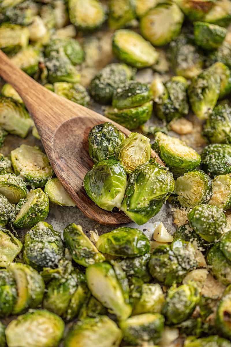 A wooden spoon scoops up Garlic Butter Roasted Brussel Sprouts from a sheet pan.