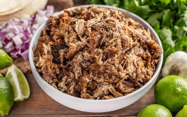 Crockpot carnitas in white bowl, surrounded by limes and corn tortillas