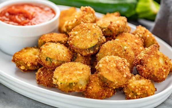 A white platter full of Fried Parmesan Zucchini Chips with a white ramekin full of marinara sauce.