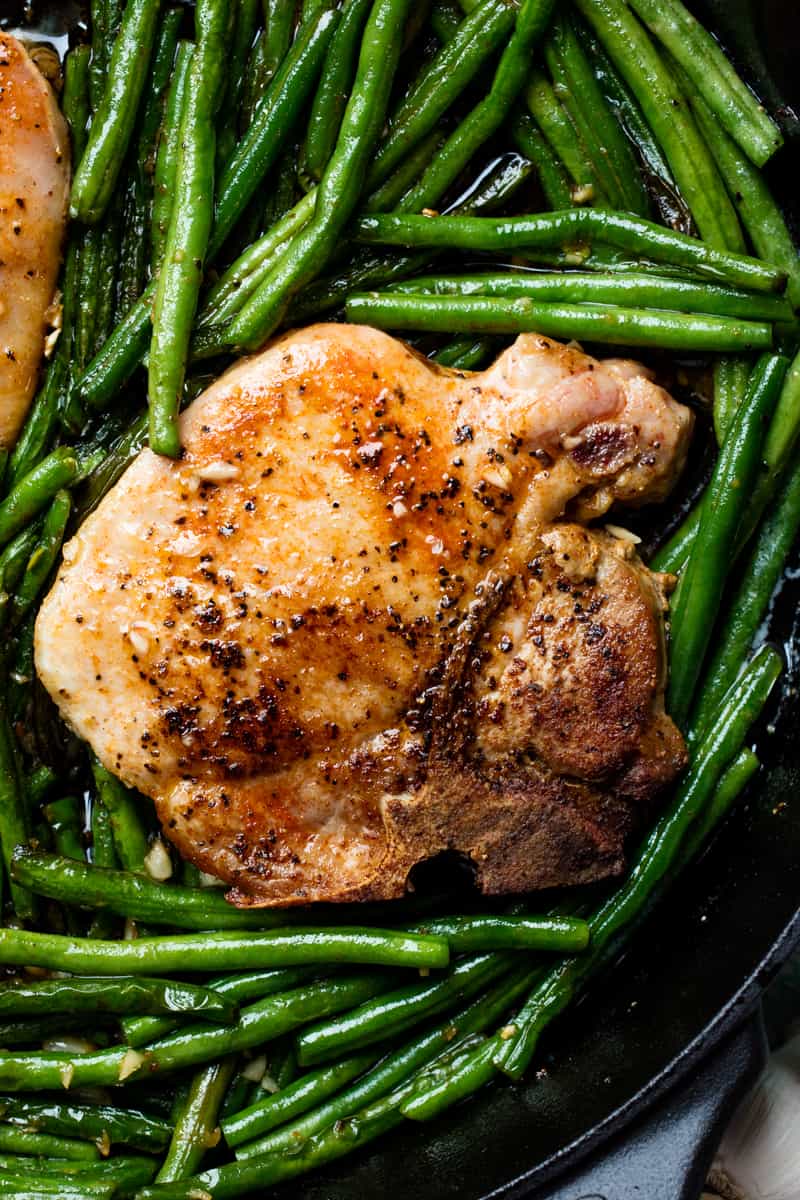 Close up of a pork chop surrounded by green beans in a cast-iron skillet.