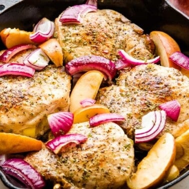 Apple stuffed pork chops in a skillet with onion slices and apple slices.