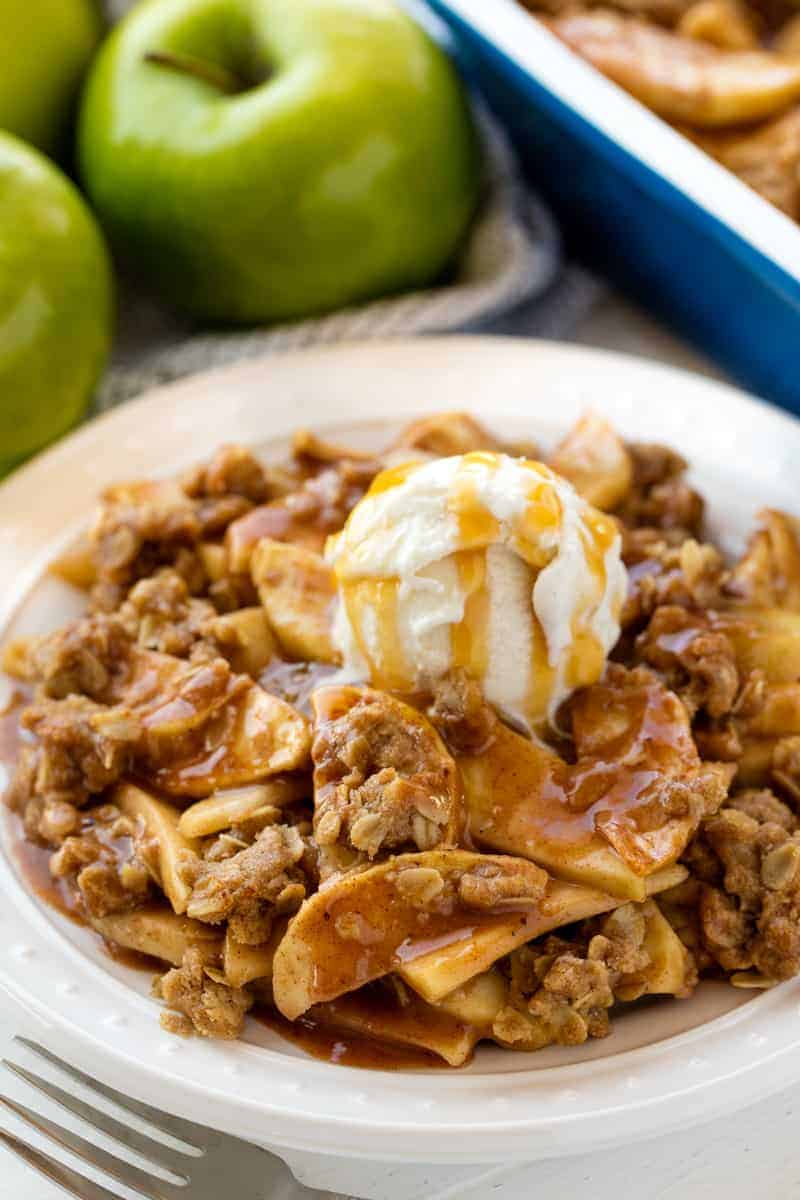 Apple Crisp served up on a white plate topped with a scoop of ice cream and caramel.