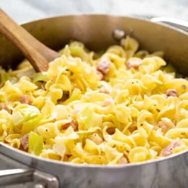 Angled view of Haluski in a stainless still pan with a wooden spoon in it.