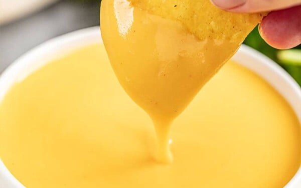 A hand dipping a tortilla chip into a small bowl of cheese sauce