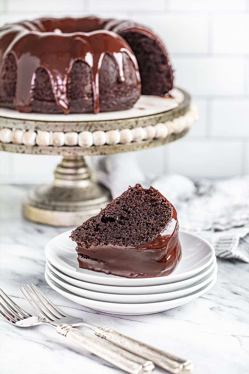 a slice of chocolate bundt cake served on a stack of small plates with the rest of the cake on a cake stand in the background