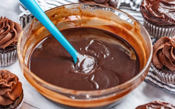 A bowl of chocolate ganache surrounded by chocolate cupcakes with ganache frosting.