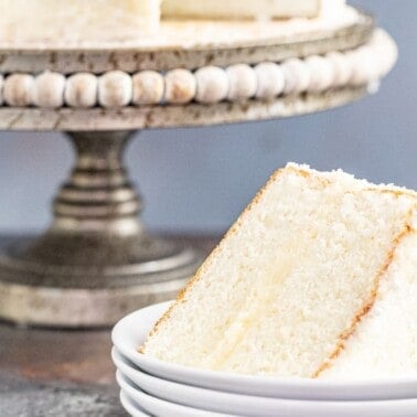 A slice of white cake on a stack of white plates with a whole white cake on a cake platter in the background.