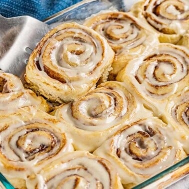 A spatula picking up a cinnamon roll from a glass baking pan.