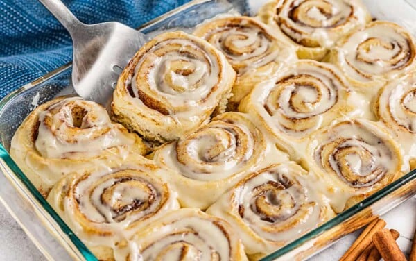 A spatula picking up a cinnamon roll from a glass baking pan.