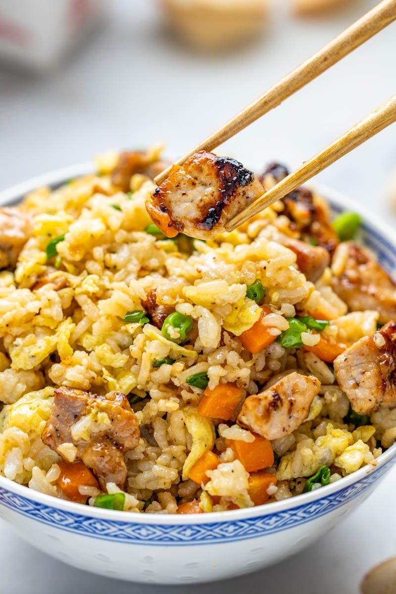 A bowl of pork fried rice with chopsticks holding a piece of pork.