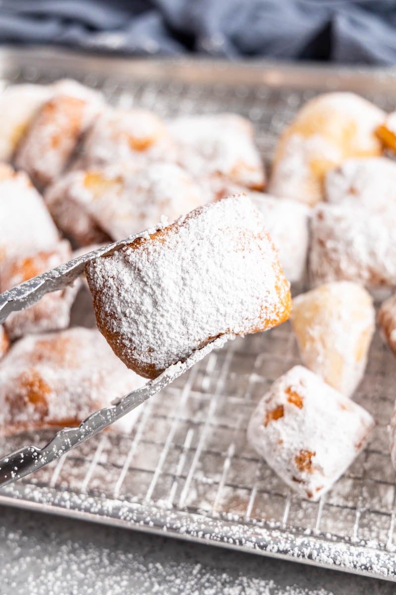 A pair of tongs holding a beignet over the baking sheet.